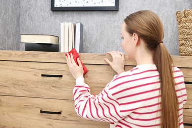 Photo of Woman cleaning wooden chest od drawers with rag at home