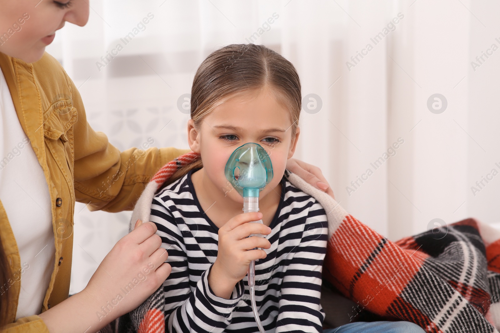 Photo of Mother covering her daughter with plaid while she using nebulizer for inhalation at home