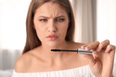 Photo of Emotional young woman holding mascara brush with fallen eyelashes indoors, closeup