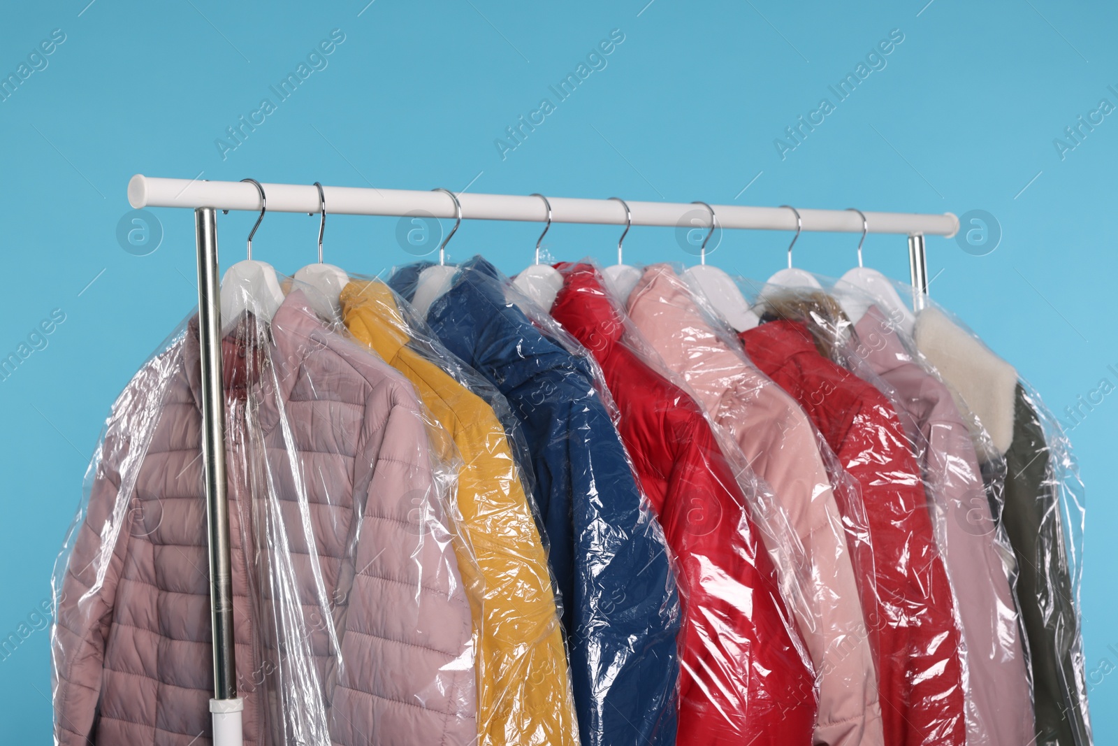 Photo of Dry-cleaning service. Many different clothes in plastic bags hanging on rack against light blue background