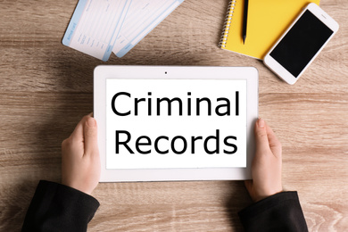 Image of Woman holding tablet with words CRIMINAL RECORDS on wooden background, top view