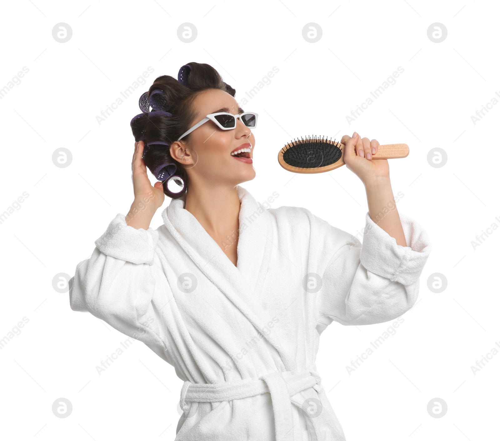 Photo of Happy young woman in bathrobe with hair curlers and sunglasses singing into hairbrush on white background