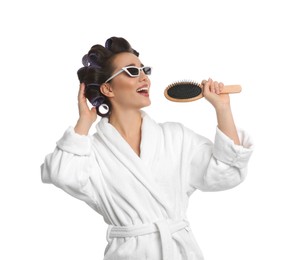 Happy young woman in bathrobe with hair curlers and sunglasses singing into hairbrush on white background