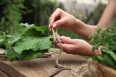 Photo of Woman tying bunch of fresh green leaves with twine at wooden table outdoors, closeup. Drying herbs