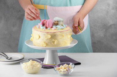 Woman putting candles on cake decorated with macarons and marshmallows at white table, closeup