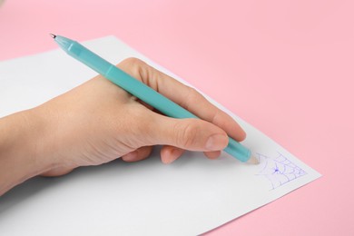 Woman erasing web drawn with erasable pen on sheet of paper against pink background, closeup