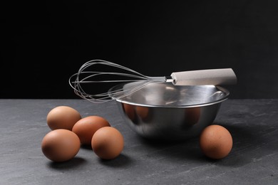 Metal whisk, raw eggs and bowl on dark grey table