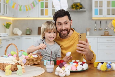Photo of Father and his cute son making selfie while painting Easter eggs at table in kitchen