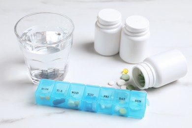 Photo of Weekly pill box with medicaments and glass of water on white marble table