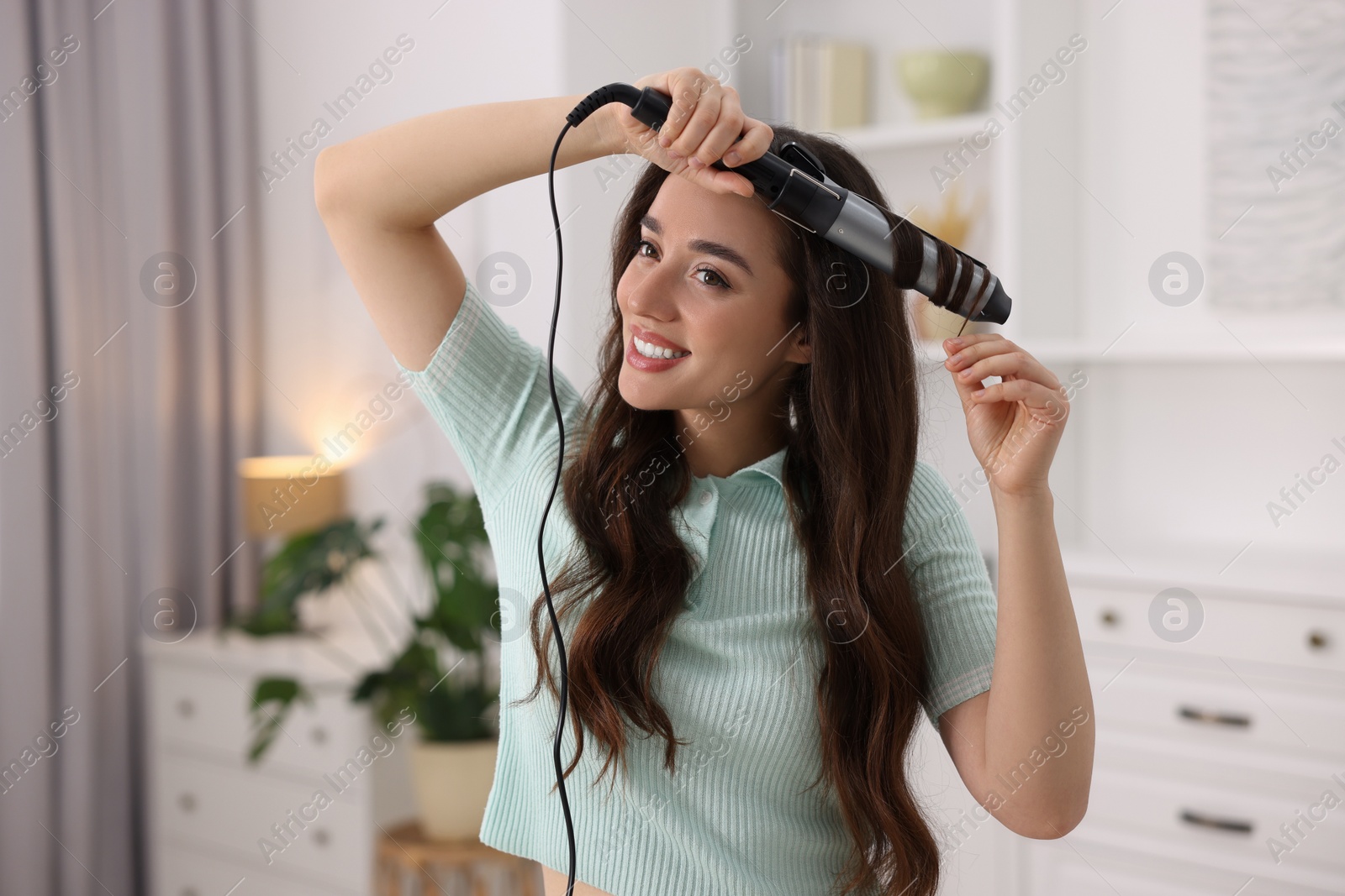 Photo of Smiling woman using curling hair iron at home