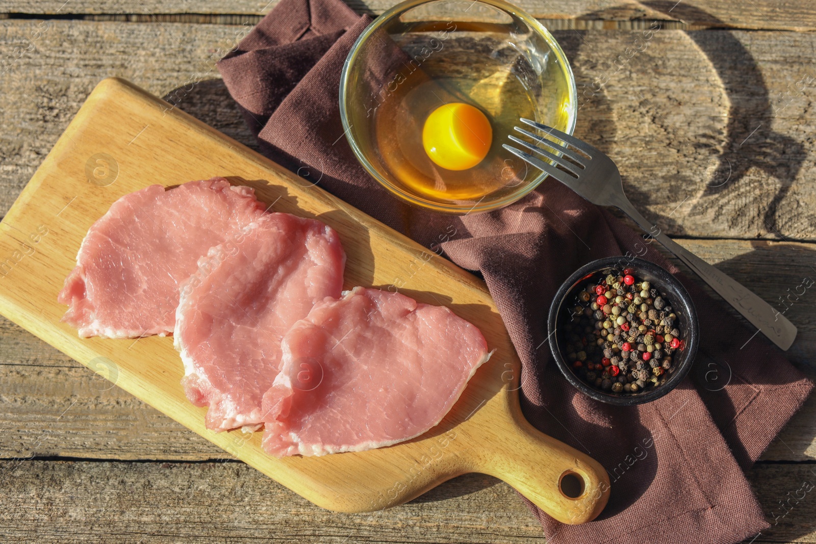 Photo of Cooking schnitzel. Raw pork slices, egg and peppercorns on wooden table, flat lay