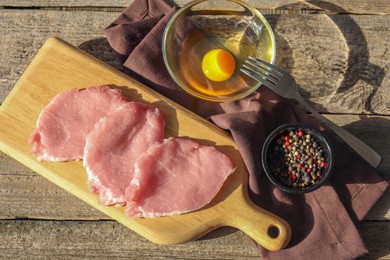 Photo of Cooking schnitzel. Raw pork slices, egg and peppercorns on wooden table, flat lay