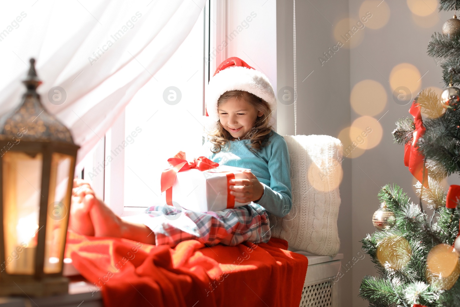 Photo of Cute little child in Santa hat with Christmas gift box sitting on windowsill at home