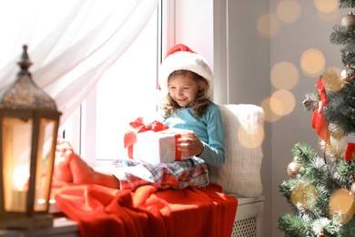 Photo of Cute little child in Santa hat with Christmas gift box sitting on windowsill at home