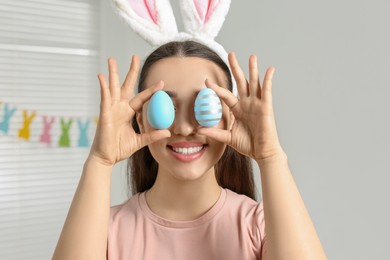 Happy woman in bunny ears headband holding painted Easter eggs near her eyes indoors