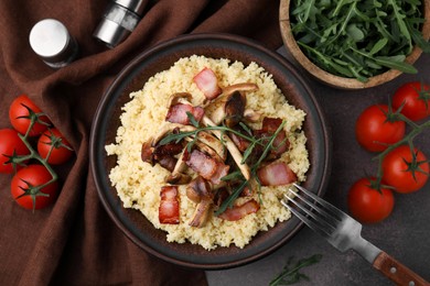 Photo of Tasty couscous with mushrooms and bacon in bowl served on brown table, flat lay