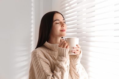 Photo of Young woman in warm sweater with cup of hot drink near window at home. Winter season