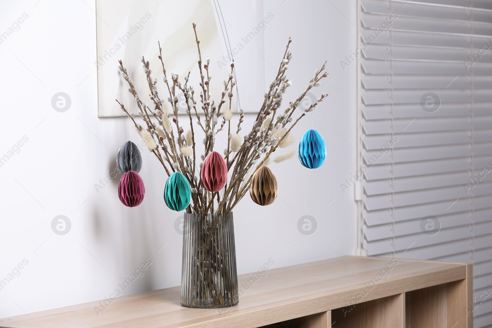 Photo of Beautiful pussy willow branches with paper eggs in vase on shelving unit at home. Easter decor