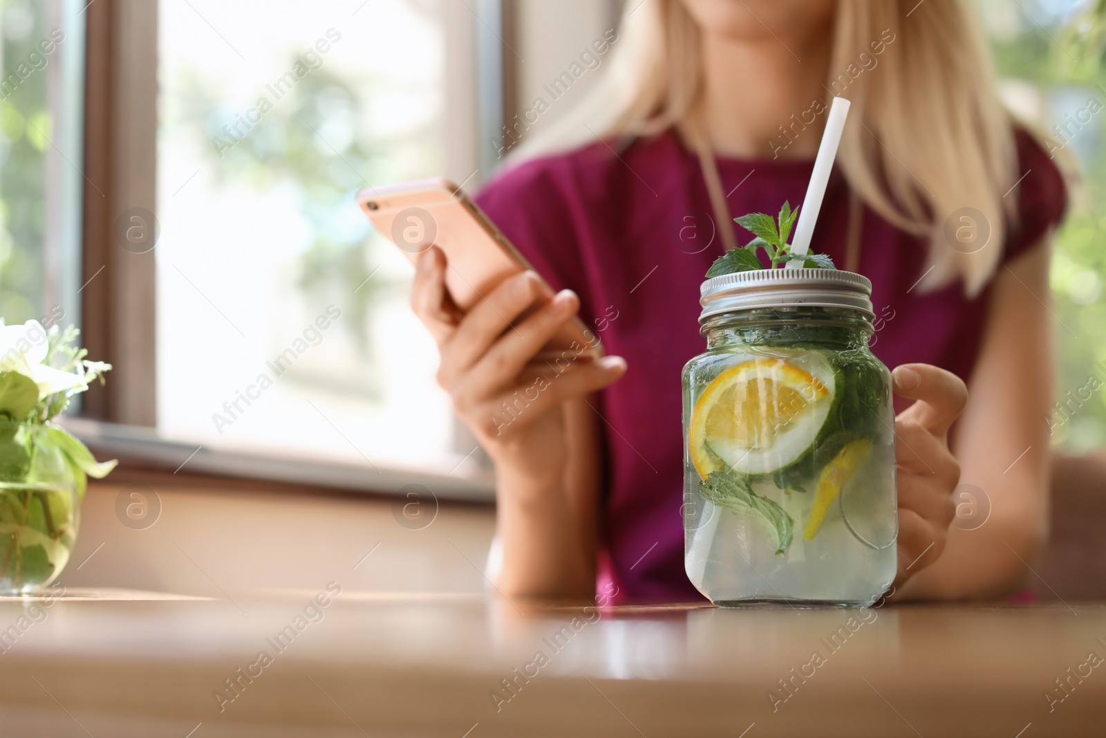 Photo of Young woman using mobile phone while drinking tasty natural lemonade at table, closeup. Detox drink