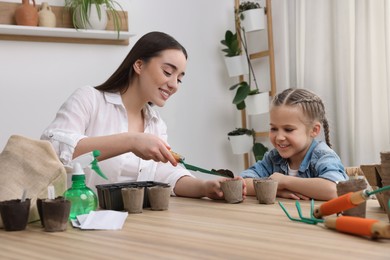 Photo of Mother showing her daughter how to fill peat pots with soil at wooden table indoors. Growing vegetable seeds