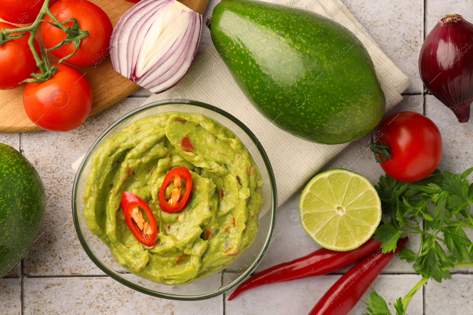 Photo of Bowl of delicious guacamole and ingredients on white tiled table, flat lay