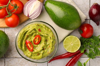 Bowl of delicious guacamole and ingredients on white tiled table, flat lay