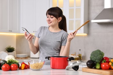 Happy young housewife with spoon using smartphone while cooking at white marble table in kitchen