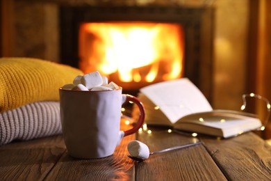 Mug with hot cocoa, marshmallows, lights and book on wooden table near fireplace