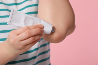 Woman applying ointment from tube onto her elbow on pink background, closeup