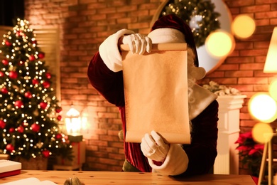 Photo of Santa Claus with blank wish list at table indoors