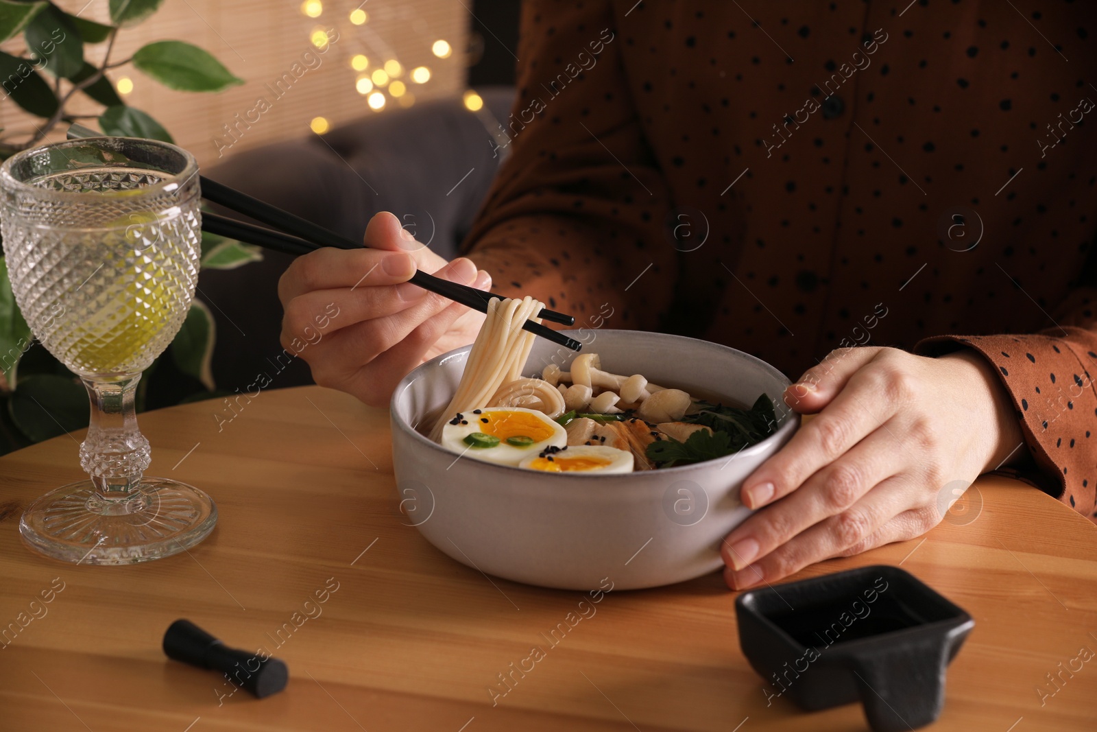 Photo of Woman eating delicious ramen with chopsticks at wooden table indoors, closeup. Noodle soup