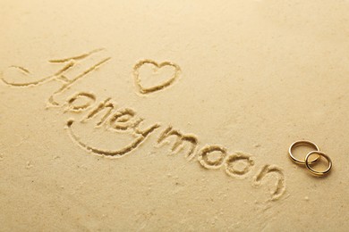 Photo of Word Honeymoon and heart written on sand and two golden rings, closeup