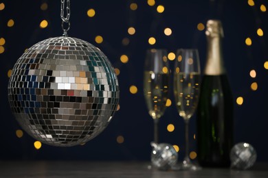 Photo of Shiny disco ball hanging over table with champagne against blurred lights, space for text