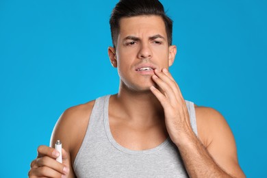 Photo of Emotional man with herpes applying cream on lips against light blue background