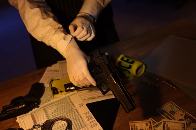 Photo of Professional detective in protective gloves examining handgun indoors at night, closeup