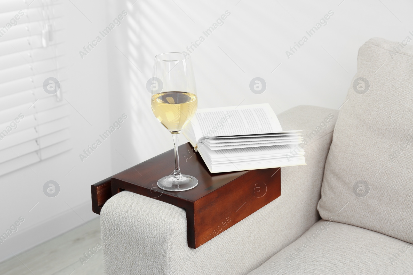 Photo of Glass of white wine and open book on sofa with wooden armrest table in room. Interior element