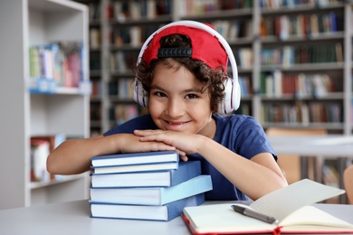 Photo of Cute little boy with headphones and books at table in library reading room