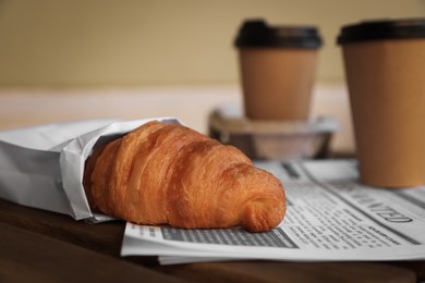 Photo of Tasty croissant, newspaper and paper cups of coffee on wooden table, closeup