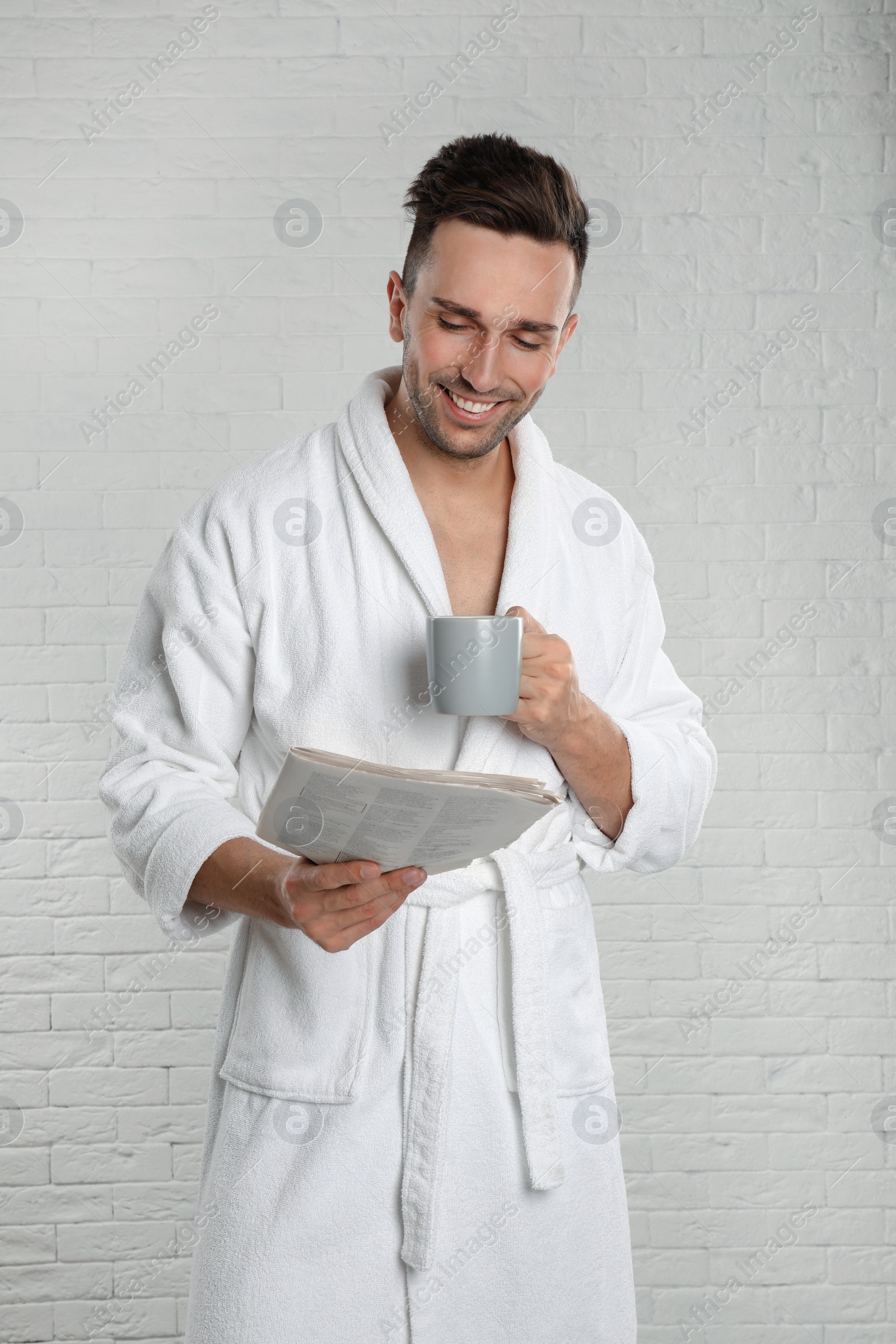 Photo of Young man in bathrobe with cup of coffee reading newspaper near white brick wall