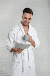 Photo of Young man in bathrobe with cup of coffee reading newspaper near white brick wall