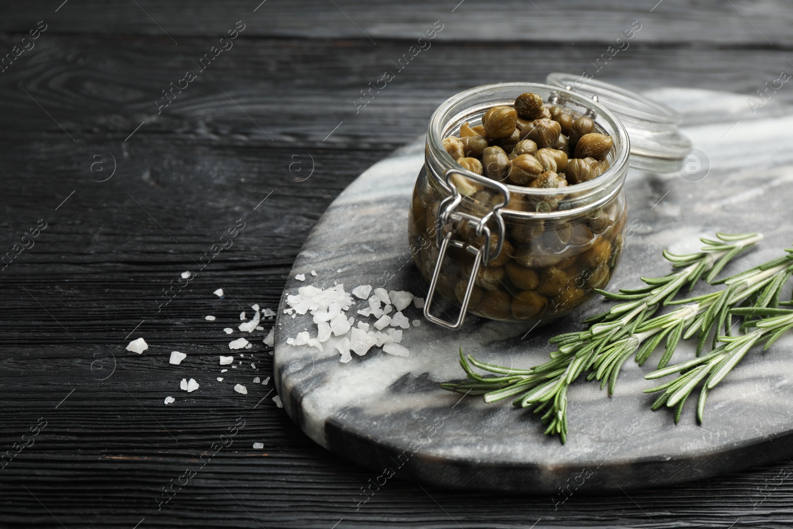 Photo of Tasty capers in glass jar, salt and rosemary on black wooden table, space for text