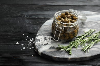 Tasty capers in glass jar, salt and rosemary on black wooden table, space for text