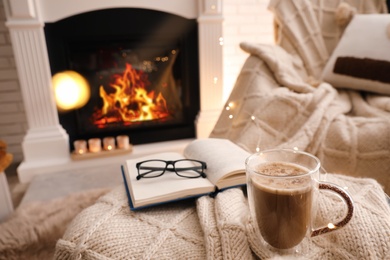 Cup of coffee, book and glasses near fireplace indoors, space for text. Cozy atmosphere