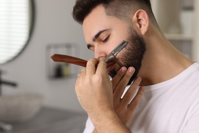 Handsome young man shaving beard with blade indoors