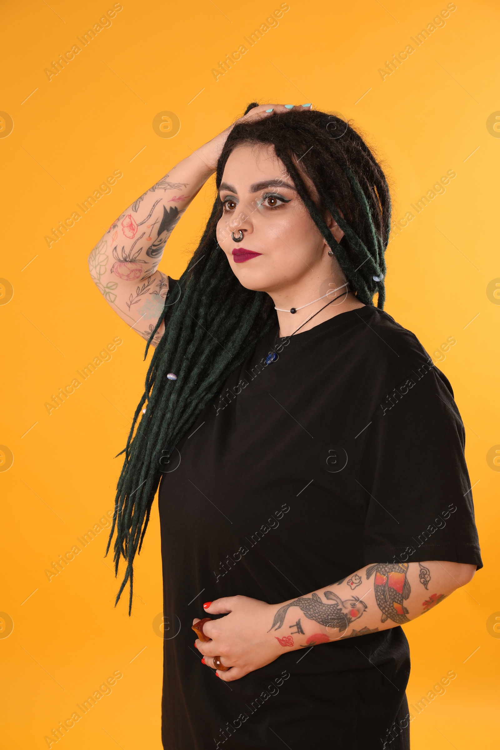 Photo of Beautiful young woman with tattoos on arms, nose piercing and dreadlocks against yellow background