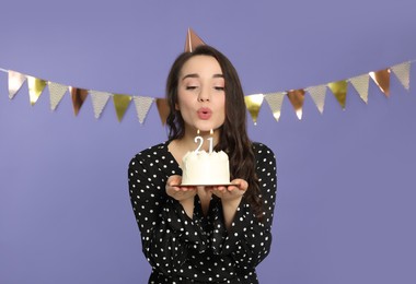 Photo of Coming of age party - 21st birthday. Woman holding delicious cake and blowing number shaped candles on violet background