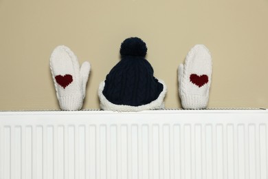Modern radiator with knitted hat and mittens near beige wall indoors
