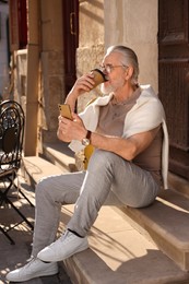 Photo of Handsome senior man sitting on doorstep with smartphone and drinking coffee outdoors