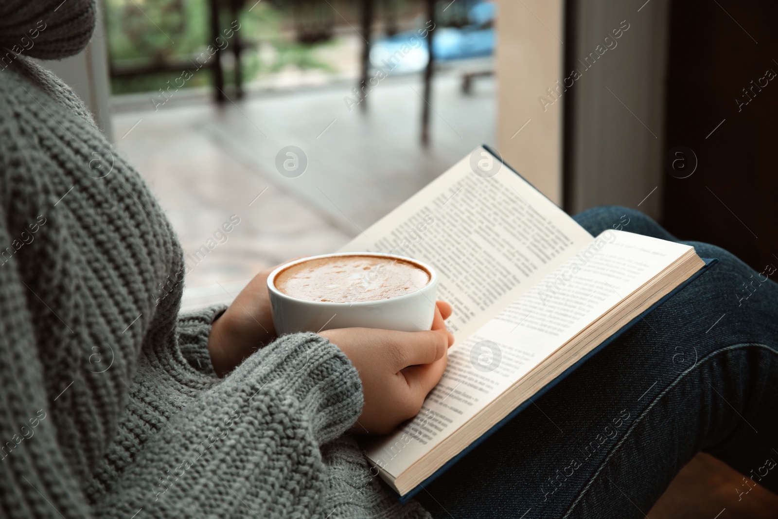 Photo of Woman with cup of coffee reading book near window indoors, closeup