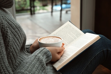 Photo of Woman with cup of coffee reading book near window indoors, closeup